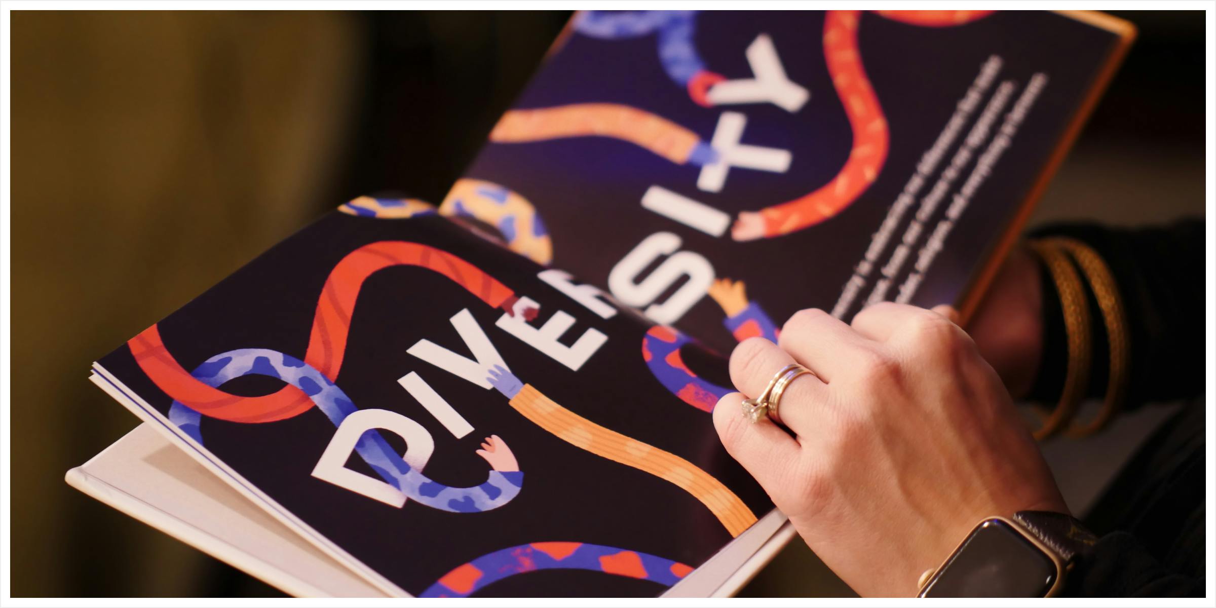 A hand holding a book, the word diversity across the spread with differnet hands composing the word